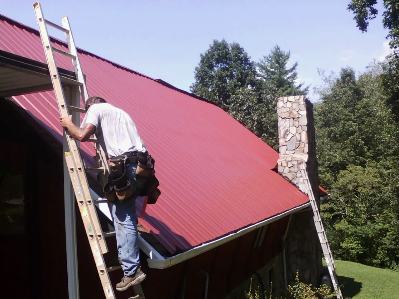 Roofer on a ladder beside a red metal roof and stone chimney — Soffit & Fascia Repair by Roofing Brothers in Selden